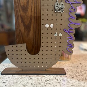 May include: A wooden earring holder in the shape of the letter "J" with multiple holes for earrings. The holder has a dark brown base and a lighter brown body. The name "Jaylyn" is written in purple script on the side. Several pairs of earrings are displayed.