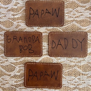 以下が含まれることがあります： 白いステッチと手書きのテキストが入った、4つの茶色の革パッチ。「PAPAW」、「GRANDPA BOB」、「DADDY」、「PAPAW」と書かれています。パッチは長方形で、手作りのようです。