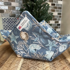 May include: A blue fabric bowl cozy with a seashell and starfish pattern. The cozy is quilted and has a label that reads "Handmade by FLPatrickProvisions" with a small American flag.