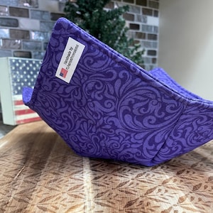 Op de afbeelding: Een paarse kom cozy met een wervelend bloemmotief. De cozy is handgemaakt en heeft een label met de tekst "Handmade by FLPatriotProvisions" met een kleine Amerikaanse vlag. De cozy staat op een houten oppervlak.