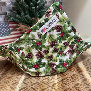 May include: A white fabric pot holder with a green and brown pine cone and holly berry pattern. The pot holder is folded into a triangle shape and has a label that reads "Handmade by FLPatriotProvisions".