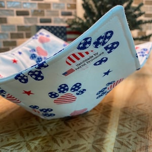 Puede incluir: Cuenco de tela blanca con un estampado patriótico de huellas de patas en rojo, blanco y azul. El cuenco presenta estrellas y rayas, con el texto "Handmade by Patriot Provisions". El cuenco está sobre una superficie de madera.