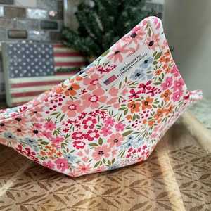 May include: A floral print bowl cozy with a white background and pink, orange, and blue flowers. The cozy is handmade and features a tag that reads "handmade by FLPatriotProvisions."