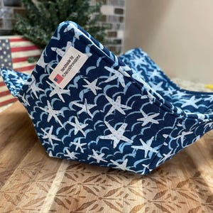 Peut inclure: Un protège-bol bleu marine avec un motif d'étoiles de mer blanches. Le protège-bol est fait à la main par F. Patrick Provisions. Il est conçu pour garder les bols au chaud et est posé sur une surface en bois.