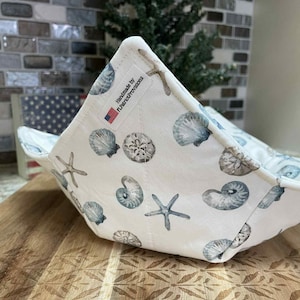 Puede incluir: Un protector de cuenco de tela blanca con un estampado de conchas marinas y estrellas de mar en tonos azules y grises. El protector está hecho a mano y presenta una etiqueta con el texto "Handmade by FLPatriotProvisions" y una bandera estadounidense.