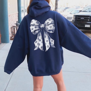 May include: A navy blue hooded sweatshirt featuring a large, centered graphic on the back. The graphic depicts a white bow with a detailed blue floral pattern throughout the ribbon. The sweatshirt has long sleeves and a hood, providing a loose fit.