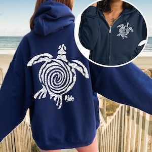 May include: A navy blue hooded sweatshirt featuring a large white graphic of a sea turtle with a spiral shell pattern on the back and the word Aloha printed below it. A circular inset shows the front of the same navy blue zip-up hoodie.