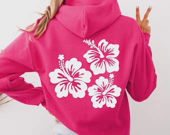 May include: A hot pink hoodie featuring a white floral design on the back. The design consists of three hibiscus flowers. The hoodie has a hood and long sleeves. The model is wearing light blue jeans.
