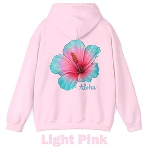 May include: A light pink hooded sweatshirt featuring a large, colorful hibiscus flower graphic on the back. The flower displays a gradient of pink, magenta, and teal blue tones. Below the flower, the word Aloha is printed in a teal script font.