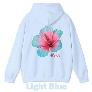 May include: A light blue hooded sweatshirt featuring a large, colorful hibiscus flower graphic on the back. The flower displays a gradient of pink and teal hues. Below the flower, the word Aloha is printed in a dark red, cursive script font.