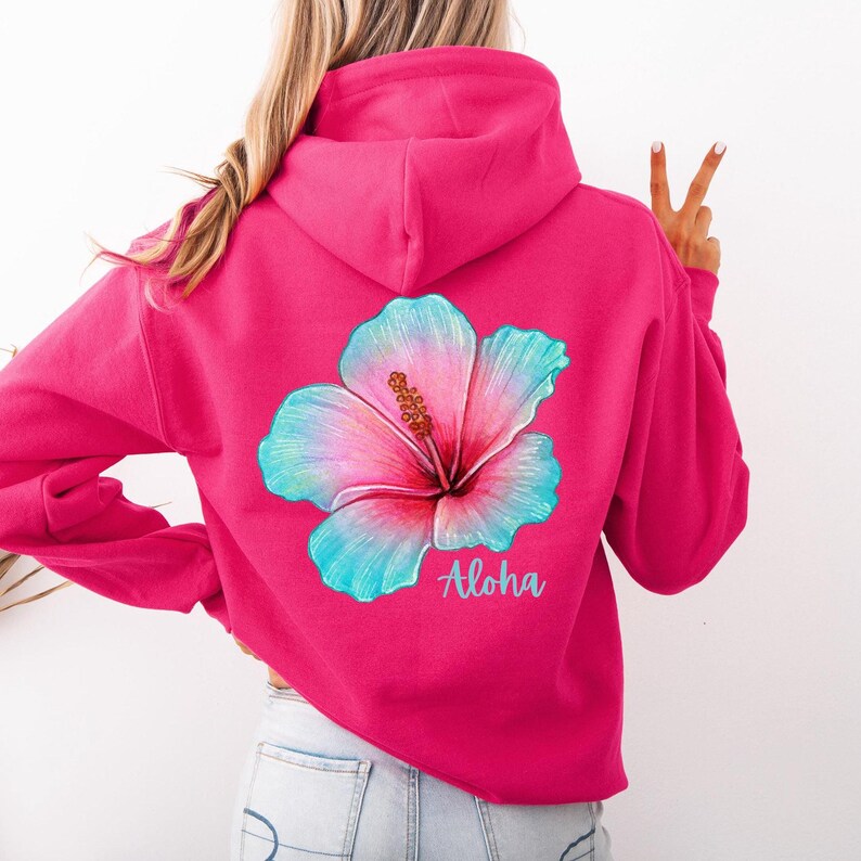 May include: A bright hot pink hooded sweatshirt featuring a large, colorful hibiscus flower graphic on the back. The flower displays a gradient of pink, blue, and teal hues. Below the floral design, the word Aloha is printed in a light blue script.