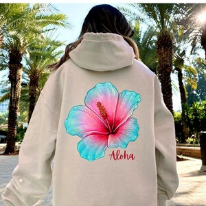 May include: A beige hooded sweatshirt featuring a large, vibrant hibiscus flower graphic on the back. The flower displays a gradient of pink and turquoise blue petals with a yellow center. The word Aloha is printed in red script below the flower.