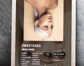 May include: A framed album cover for the album "Sweetener" by Ariana Grande. The album cover features a close-up of Ariana Grande's face, with her hair styled in a white blonde updo. The album title and artist name are printed in white text on a black background. The tracklist is printed in white text on a gray background. The album cover also includes a barcode and a Spotify logo.