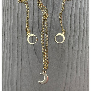 May include: A gold chain necklace with a crescent moon pendant and two circular charms. The necklace is on a gray wooden background.