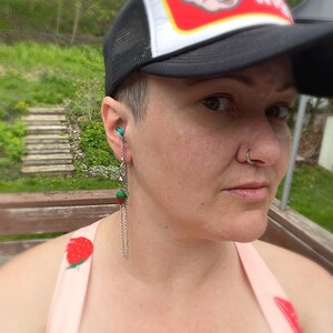 May include: A person wearing a black and white trucker hat with a red logo that says "Pig Wig". The person is wearing a pink tank top with a strawberry print. The person has a nose ring and a green and silver dangle earring.