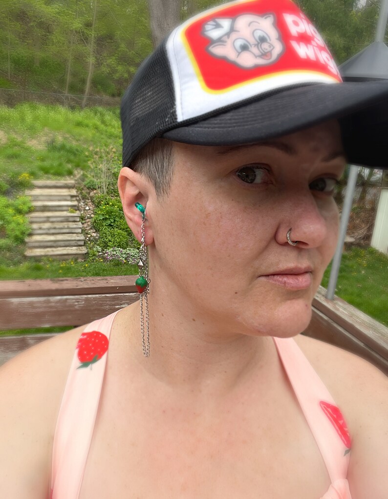 May include: A person wearing a black and white trucker hat with a red logo that says "Pig Wig". The person is wearing a pink tank top with a strawberry print. The person has a nose ring and a green and silver dangle earring.