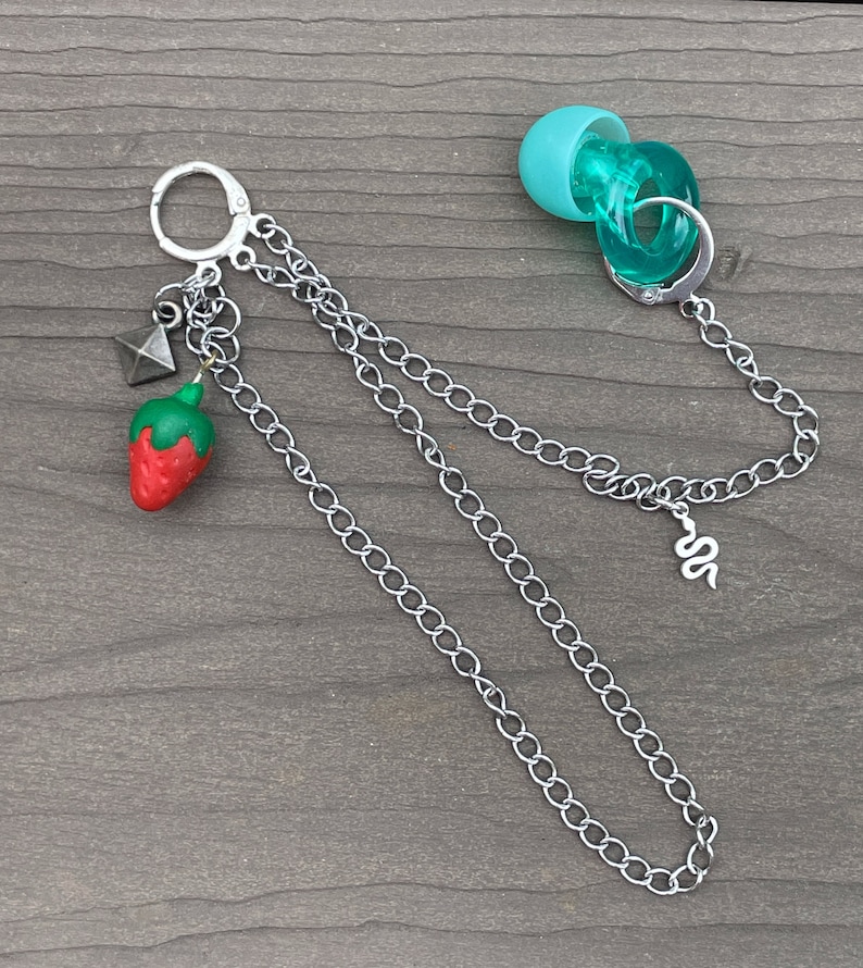 May include: A silver chain necklace with a charm of a red strawberry, a silver pyramid, and a silver snake. The necklace also has a turquoise blue charm with a silver ring.