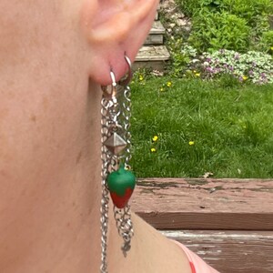 May include: A silver chain earring with a green and red strawberry charm. The earring is hanging from a person's ear.