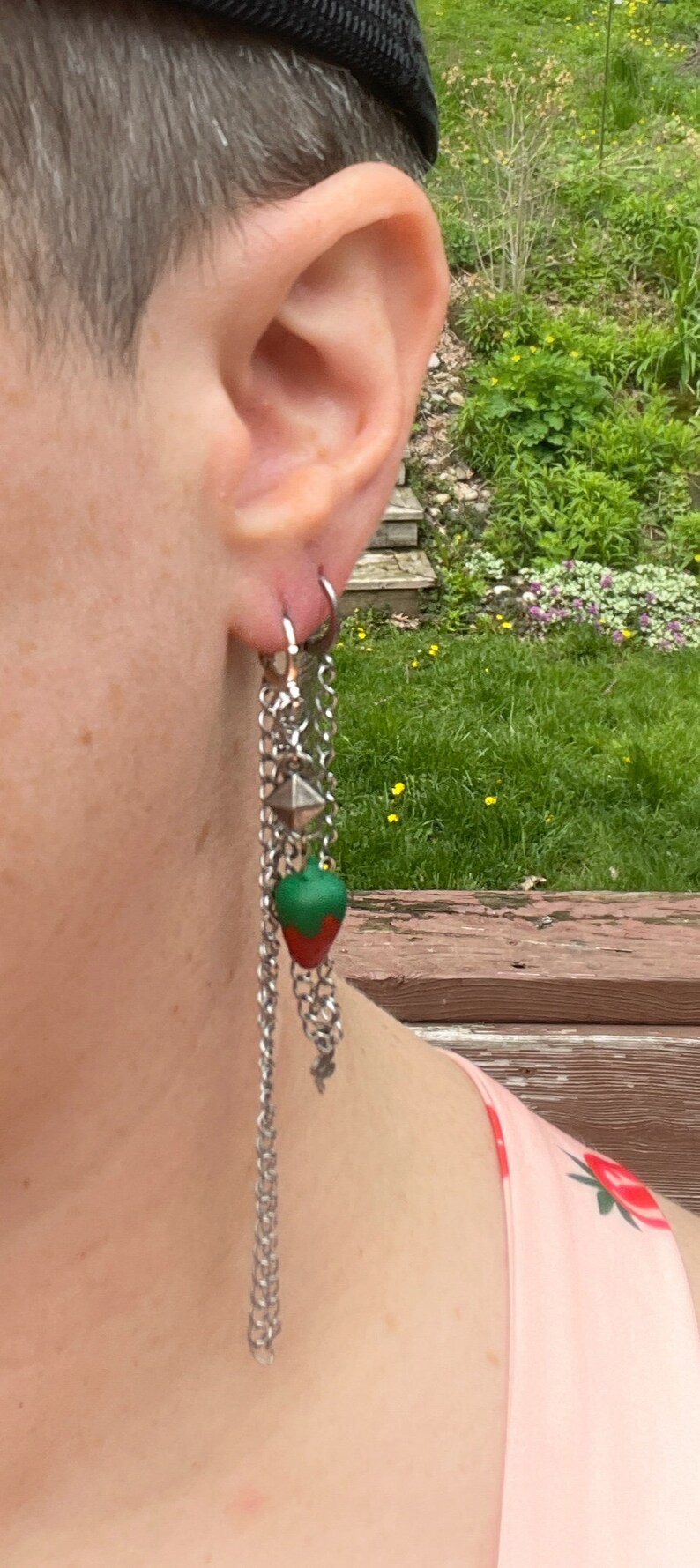 May include: A silver chain earring with a green and red strawberry charm. The earring is hanging from a person's ear.