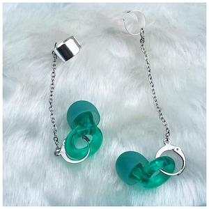 Puede incluir: Un par de pendientes verdes y plateados. Los pendientes están hechos de plástico verde retorcido y tienen una cadena plateada unida a un brazalete plateado. El brazalete está diseñado para ser usado en la oreja.