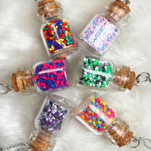 May include: Six glass bottle necklaces filled with colorful sprinkles. Each bottle has a cork stopper and hangs from a silver chain. The sprinkles are in various colors, including red, blue, green, and purple.