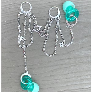 Puede incluir: Un par de pendientes de aro plateados con cadenas y colgantes. Los colgantes incluyen estrellas, planetas y pequeñas cuentas plateadas. Los pendientes están unidos a colgantes de plástico turquesa en forma de chupete.