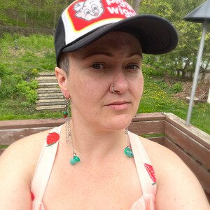 May include: A person wearing a black and white trucker hat with the words "Piggly Wiggly" on the front. They are wearing a pink dress with a strawberry pattern and have long, dangling earrings with green beads.