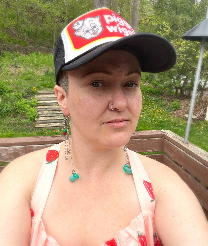 May include: A person wearing a black and white trucker hat with the words "Piggly Wiggly" on the front. They are wearing a pink dress with a strawberry pattern and have long, dangling earrings with green beads.