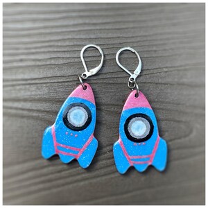 May include: A pair of blue and pink rocket ship earrings with silver hooks. The rockets have a white and black circle in the center and pink details.