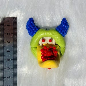 Puede incluir: Un colgante con forma de monstruo que presenta una boca verde lima, dos cuernos azules texturizados, colmillos blancos y una lengua amarilla cubierta de esferas rojas. Una regla metálica junto al objeto muestra una altura de 6,5 centímetros.