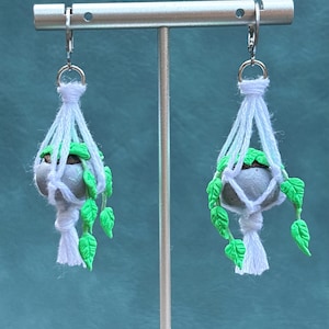 May include: A pair of handmade earrings featuring miniature gray planters suspended in white macrame. Each planter holds green foliage and is attached to a silver-tone ear wire. The earrings are displayed on a silver stand against a teal background.