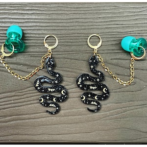 May include: A pair of earrings with a black snake design, adorned with white stars and crescent moons. They are linked to teal plugs by gold chains. The earrings are displayed on a wooden surface.