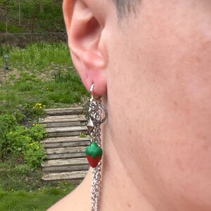 May include: A silver chain earring with a red and green strawberry charm. The earring is hanging from a person's ear.