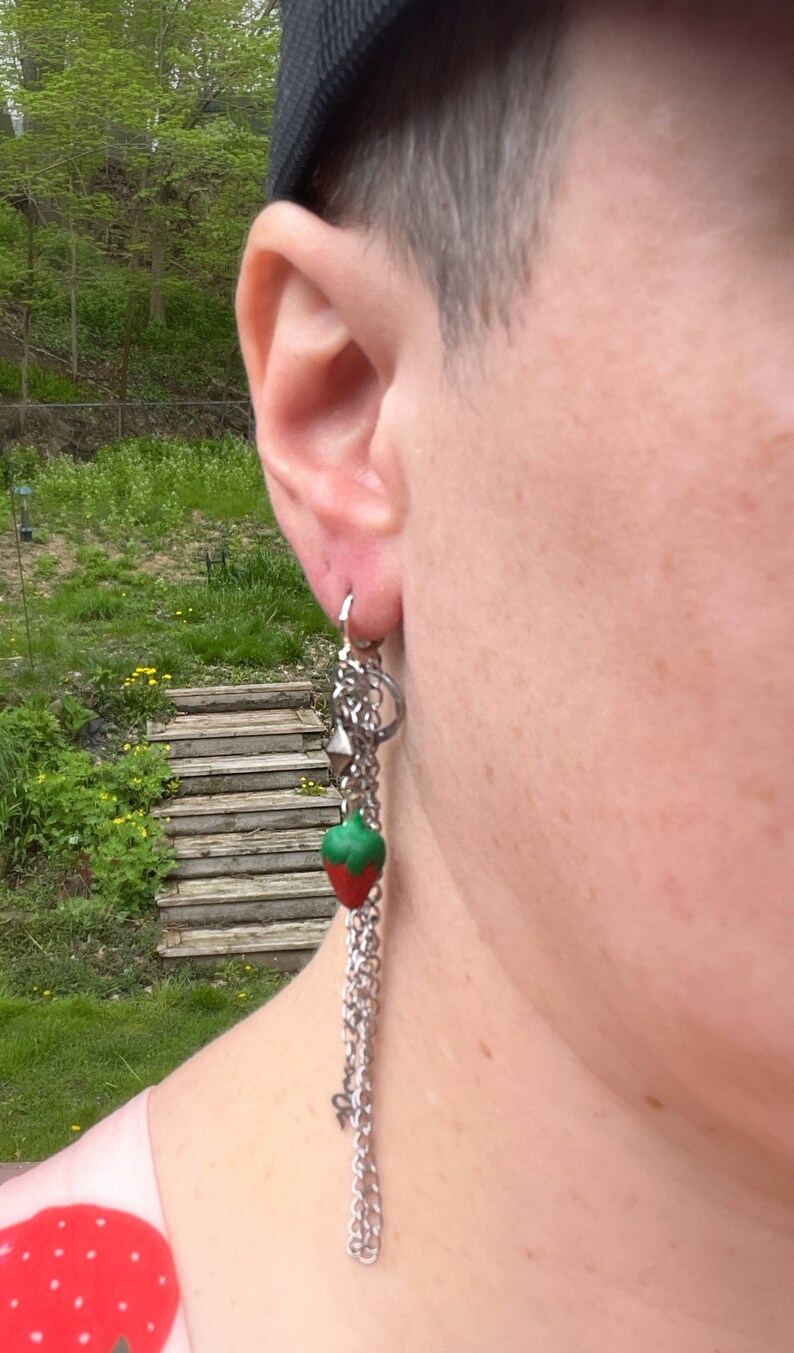 May include: A silver chain earring with a red and green strawberry charm. The earring is hanging from a person's ear.