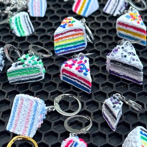May include: A collection of miniature cake slice keychains in various colors, including rainbow, pink, and purple. Each keychain features a detailed cake slice design with sprinkles and frosting, attached to a silver or gold keyring.