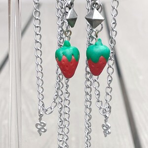 May include: A pair of silver chain earrings with a strawberry charm on each earring. The earrings are hanging from a clear acrylic display stand.
