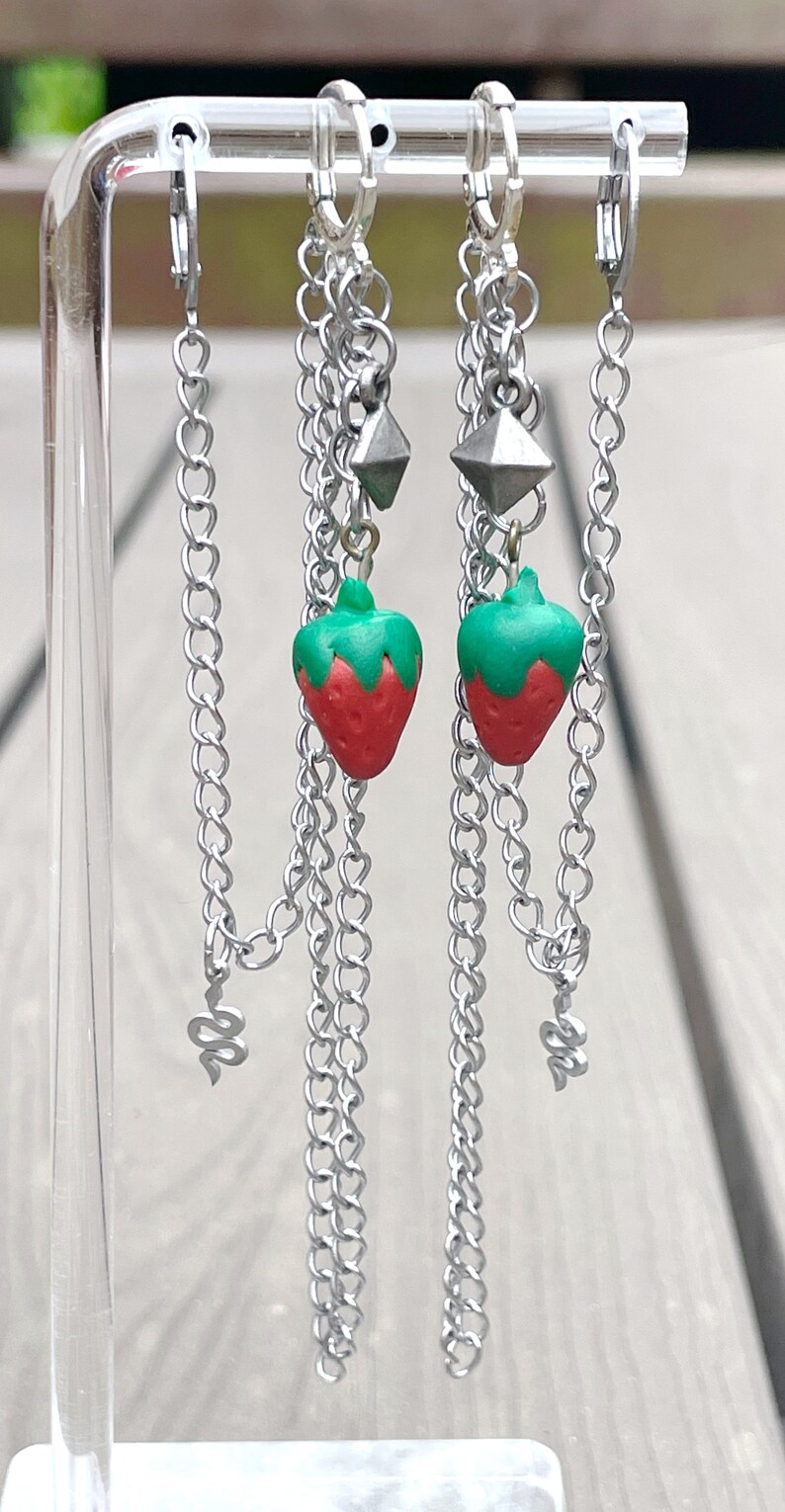 May include: A pair of silver chain earrings with a strawberry charm on each earring. The earrings are hanging from a clear acrylic display stand.