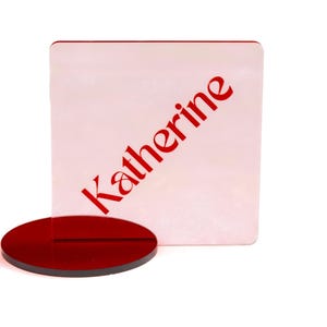May include: A red acrylic place card holder with the name "Katherine" printed in red. The holder has a square card and a round base, both in red. The background is white.