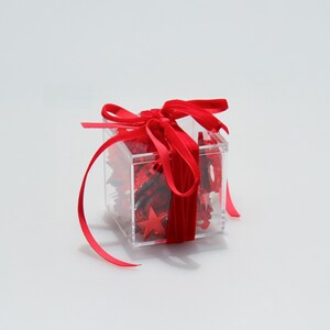 May include: A clear, square gift box tied with a vibrant red satin ribbon. The box is filled with red star-shaped confetti and decorative filler. The ribbon is tied in a bow on top, with long flowing tails.