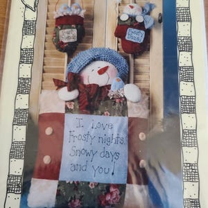 May include: A craft pattern titled "Luv 'N Stuff" featuring a snowman and two hanging ornaments. The snowman wears a blue hat and has a red nose. The ornaments say "Best Friends" and "Frosty Daze". The text on the quilt reads "I love frosty nights. Snowy days and you!"