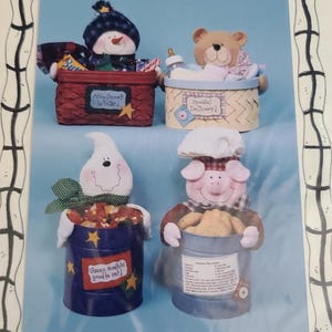 May include: Four whimsical, handcrafted container decorations. A snowman in a red basket, a teddy bear in a beige basket, a ghost in a blue container, and a pig in a blue container. Each container has a unique label with text.