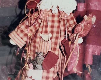 May include: A handmade Santa Claus doll with a red and white plaid coat, white beard, and a pocket with a star design. The doll has a bird applique and holds a small rocking horse. Decorative elements include a small bell and a sprig of greenery.