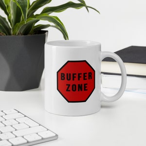 Puede incluir: Taza de cerámica blanca con un letrero octogonal rojo que dice "BUFFER ZONE" en letras negras. La taza tiene un asa curva y está sobre una superficie blanca. Una planta en una maceta gris oscuro y libros están en el fondo.