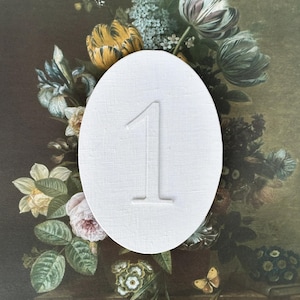 May include: An oval-shaped white card featuring a textured linen-like finish with the number 1 embossed in the center. The card is positioned against a dark background displaying a detailed floral arrangement with various colorful flowers and butterflies.