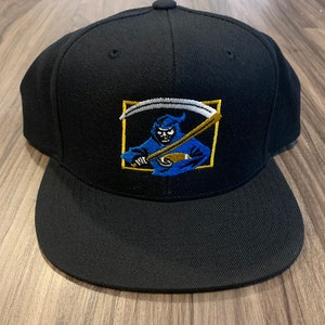 Könnte beinhalten: Eine schwarze, passgenaue Baseballkappe mit einem gestickten Bild eines blauen Sensenmanns, der eine Sense in einem goldenen Quadrat hält.