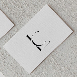 Puede incluir: Dos tarjetas de visita blancas con letras negras. Las letras "LC" están estilizadas con la "C" formando un círculo alrededor de la "L". Las tarjetas están sobre una superficie texturizada de color gris claro.