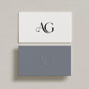 May include: Two rectangular business cards, one white and one grey. The white card has the letters "AG" in black script. The grey card has the same letters embossed. Both cards are on a light grey surface.