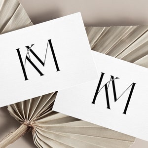 Op de afbeelding: Twee witte visitekaartjes met de initialen "KM" in zwarte letters op een beige achtergrond. De kaartjes liggen op twee gedroogde palmbladeren.