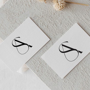 以下が含まれることがあります： 白い名刺2枚に、黒いスタイリッシュな文字「S」が書かれている。名刺は薄いグレーのざらざらした表面の上に置かれている。