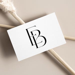 May include: A white business card with the letters "FB" in a black, elegant font. The card rests on a beige surface, with dried ornamental grass stems in the background. The overall aesthetic is minimalist and sophisticated.